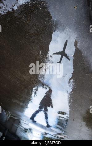 Verschwommene Silhouette der Spiegelung einer Person, die an regnerischen Tagen allein auf dem nassen Bürgersteig der Stadt läuft. Das Flugzeug fliegt über den Himmel. Abstrakte Fotografie. Stockfoto