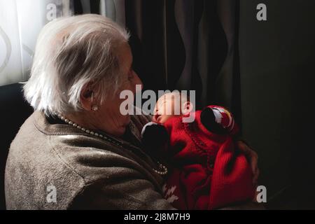 Eine alte weißhaarige Urgroßmutter hält ihr neugeborenes Enkelkind in der Hand Stockfoto