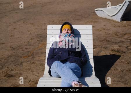 Kaukasische junge Frau in warmen Kleidern, die sich auf der Sonnenliege am Strand ausruhen Stockfoto