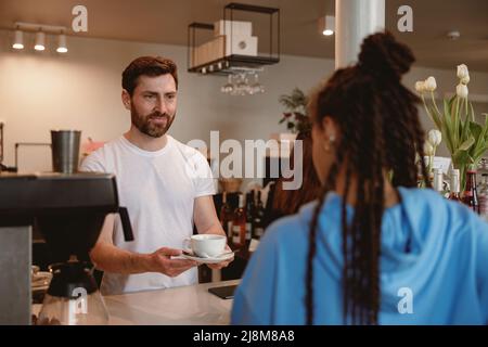 Fröhlicher, gutaussehender Kellner, der weiblichen Gästen an der Bar Kaffee serviert. Stockfoto