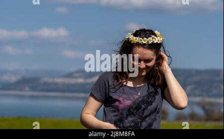 Selektive Fokusaufnahme einer wellenförmigen Frau mit Gänseblümchen-Krone auf ihrem Kopf, die lächelt, während sie nach unten blickt. Stockfoto