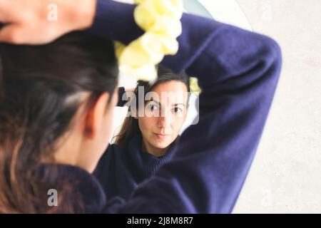 Junge attraktive Frau, die sich im Spiegel ansieht und ihr Haar mit einem Scrunchie zu einem Pferdeschwanz bindet Stockfoto