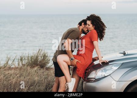 Junges Paar auf Roadtrip, Mann intim Küssen Frau in den Hals auf der Haube ihres SUV-Autos auf dem Hintergrund des Meeres bei Sonnenuntergang Stockfoto