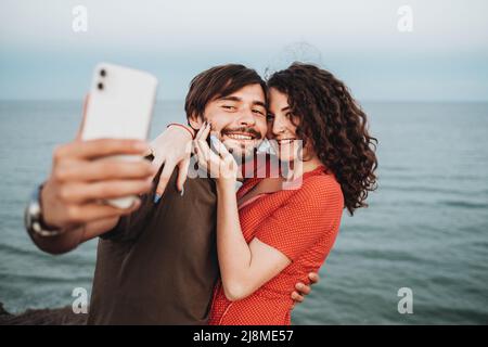 Glückliches kaukasisches Paar Mann und lockige Brünette Frau umarmen, während sie gegen das Meer stehen und Selfie auf dem Smartphone machen Stockfoto