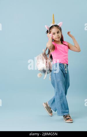 Vibrierende Tween Mädchen in rosa Hemd und Einhorn Haarband sprechen auf ihrem Telefon. Sie hat Haarsträhnen gefärbt. Posiert auf blauem Hintergrund. Stockfoto