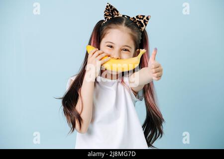 Positiv aussehendes Mädchen mit Katzenohren, die Banane als Lächeln über ihren Mund halten. Sie hat Haarsträhnen gefärbt. Posiert auf blauem Hintergrund. Stockfoto