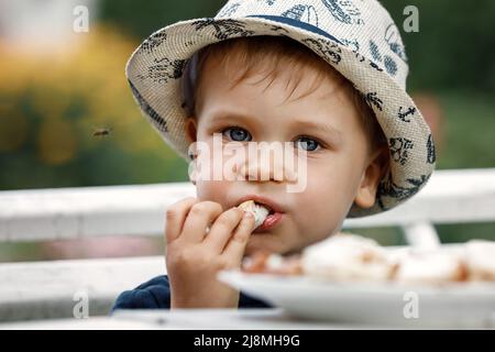 Kleiner Junge, der auf grünem Gartenhintergrund geröstetes Brot zum Frühstück isst. Gegen das Futter fliegt Hornisse. Kindheit, Nahrung, Pflege und Gesundheit Stockfoto