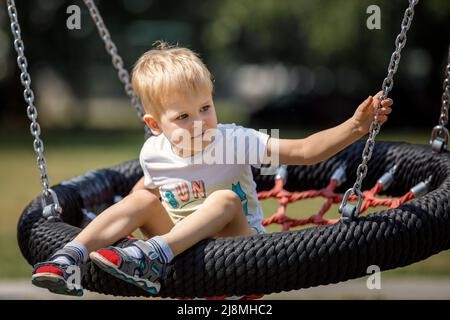 Ein fröhlicher kleiner Junge schaukelt im Park auf einer schwingenden Seilschaukel. Runder Sitz in Schwarz und Rot für die Kinderschaukel. Stockfoto