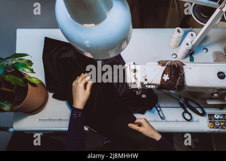 Eine Schneiderin näht Kleidung - eine modische Jacke aus Samtstoff auf einer Nähmaschine in ihrer Werkstatthalle, Draufsicht Stockfoto