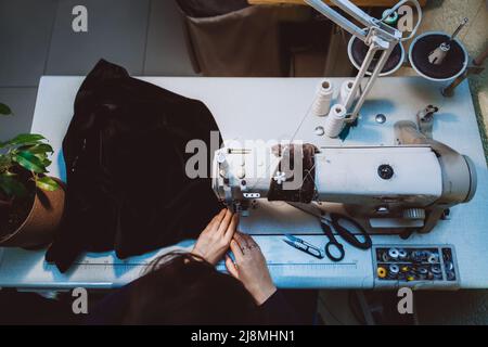 Eine Schneiderin näht Kleidung - eine modische Jacke aus Samtstoff auf einer Nähmaschine in ihrer Werkstatthalle, Draufsicht Stockfoto