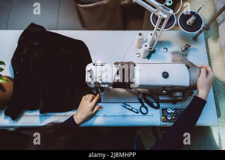 Eine Schneiderin näht Kleidung - eine modische Jacke aus Samtstoff auf einer Nähmaschine in ihrer Werkstatthalle, Draufsicht Stockfoto