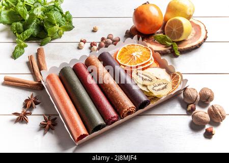 Obstpastilien verschiedener Geschmacksrichtungen und Fruchtchips oder Trockenobstscheiben in einer Geschenkbox, umgeben von frischen Früchten, Nüssen und Gewürzen und frischem Basilikum Stockfoto