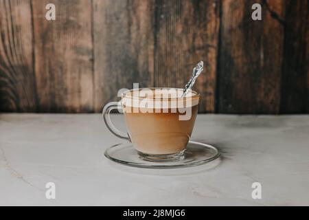 Kaffee mit Milch in einer transparenten Tasse mit einem eingeprägten Löffel auf einem Teller auf einem dunklen Holzhintergrund Stockfoto