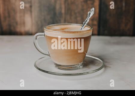 Kaffee mit Milch in einer transparenten Tasse mit einem eingeprägten Löffel auf einem Teller auf einem dunklen Holzhintergrund Stockfoto