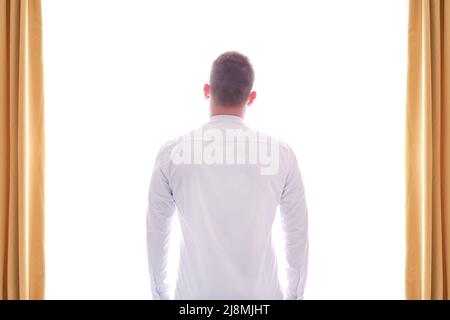 Ein Mann in einem weißen Hemd steht mit seinem Rücken und Blick aus dem Fenster mit hellem weißem Licht und goldenen Vorhängen. Licht der Zukunft Stockfoto