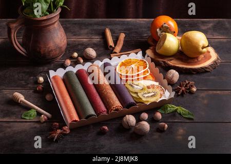 Obstpastilien verschiedener Geschmacksrichtungen und Fruchtchips oder Trockenobstscheiben in einer Geschenkbox, umgeben von frischen Früchten, Nüssen und Gewürzen und frischem Basilikum Stockfoto