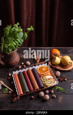 Obstpastilien verschiedener Geschmacksrichtungen und Fruchtchips oder Trockenobstscheiben in einer Geschenkbox, umgeben von frischen Früchten, Nüssen und Gewürzen und frischem Basilikum Stockfoto