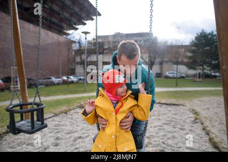 Vater schiebt seine kleine Tochter mit Down-Syndrom auf Schaukel im Freien auf dem Spielplatz. Stockfoto