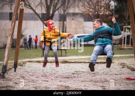 Vater mit seiner kleinen Tochter mit Down-Syndrom auf Schaukeln im Freien auf dem Spielplatz. Stockfoto