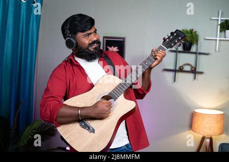 Fröhlicher junger Bartmusiker, der zu Hause Gitarre spielt - Konzept für Hobby, Entspannung am Wochenende und Freizeitaktivitäten. Stockfoto