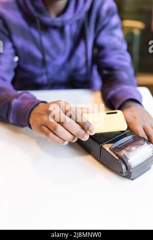 Anonymer afroamerikanischer Klient mit hohem Erntewinkel, der NFC-Technologie für kontaktloses Bezahlen mit Mobiltelefon am Terminal im Café verwendet Stockfoto