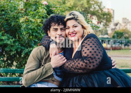 Ein Mann mit Lockenwicklern und einer Frau, die kaukasische Argentinier in die Größe nimmt, glückliche Freunde umarmt im Park, die auf einer Holzbank sitzen und lächelnd die Kamera betrachten Stockfoto