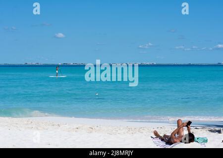 Ein junger Mann, der auf einem Brett in der Nähe des Strandes paddelt, eine Frau, die mit ihrem Smartphone auf dem Sand liegt, in Mexiko Stockfoto