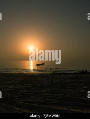 Schöner Frühmorgen bei Sonnenaufgang mit dem dunklen Strand im Vordergrund und der Sonne mit einer Silhouette eines Fischerbootes im Hintergrund. Stockfoto