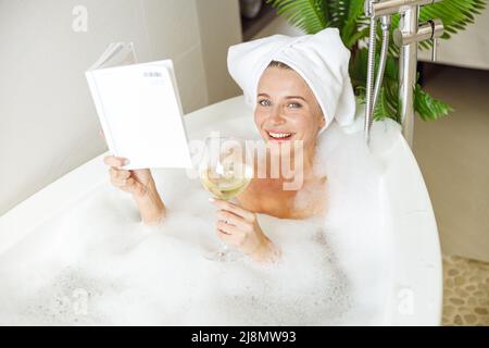 Porträt einer hübsch lächelten jungen Frau, die mit Schaum im Bad liegt, liest und Wein trinkt. Stockfoto