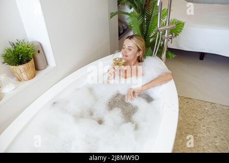 Blonde junge attraktive Frau, die in Blasen in der Badewanne liegt und Weißwein trinkt. Stockfoto