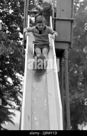 1970s, historisch, ein kleiner Junge, der eine Metallrutsche der Ära hinuntergeht, auf seinem Boden, die Füße zuerst, kleine Schwester an der Spitze, die auf ihre Wende wartet, England, Großbritannien. Stockfoto