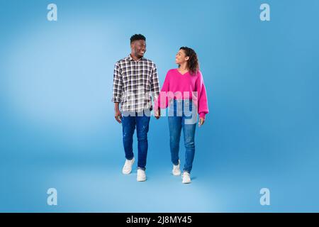 Glückliches, tausendjähriges schwarzes Paar, das die Hände hält und sich auf blauem Studiohintergrund in voller Länge anschaut Stockfoto