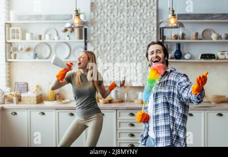 Glücklicher aufgeregt Mann mit Stoppeln in Gummihandschuhen und Frau mit Spray singen auf imaginäres Mikrofon Stockfoto