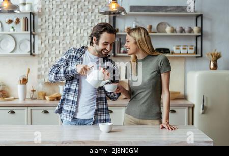 Lächelnder europäischer Millennials-Mann mit Stoppeln gießt Tee auf eine Tasse blonde Frau in einer hellen Kücheneinrichtung Stockfoto