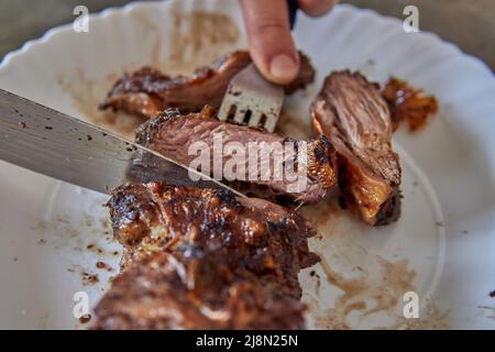 Argentinischer Grill. Menschenhände schneiden ein Stück Fleisch mit Messer und Gabel. Nahaufnahme von gegrilltem Rindersteak in Scheiben auf einem Teller Stockfoto