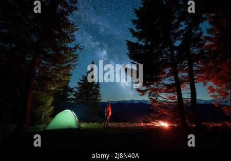 Seitenansicht des männlichen Wanderers, der aufschaut bewundern Sie die Schönheit des malerischen Sternenhimmels mit Milchstraße während der Nacht auf dem Campingplatz, Backpacker, die Berge mit Zeltlager in der Nähe des Fichtenwaldes im Nationalpark besuchen Stockfoto