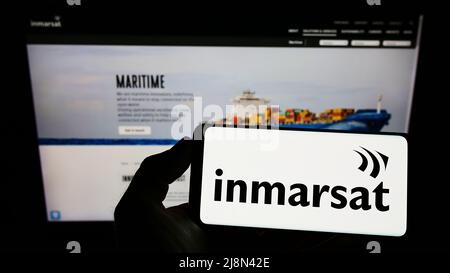 Person, die Mobiltelefon mit dem Logo des Satellitenunternehmens Inmarsat Global Limited auf dem Bildschirm vor der Business-Webseite hält. Konzentrieren Sie sich auf die Telefonanzeige. Stockfoto