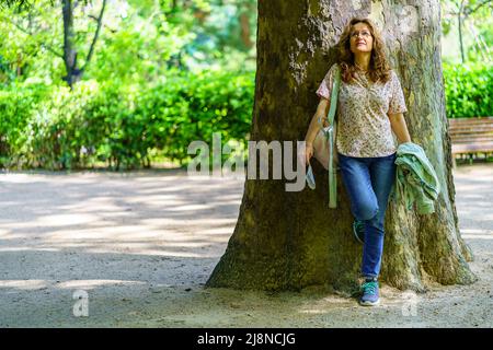Ältere Frau, die sich im Licht der warmen Frühlingssonne auf den Stamm eines riesigen Baumes lehnt. Stockfoto