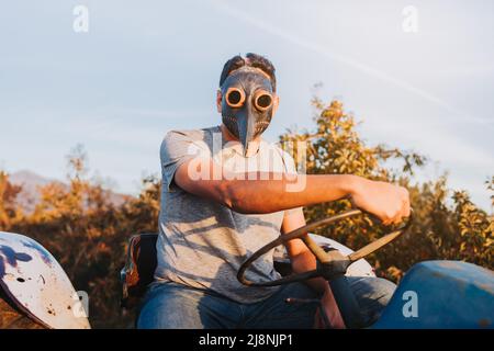 Bauer Mann mit der Pestmaske an, der mitten in seinem Ackerland einen alten Traktor fährt. Stockfoto