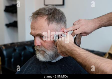 Ein alter Mann, der in einem Friseurladen einen Haarschnitt von einem Meister genießt. Ein alter Mann bekommt einen stylischen Haarschnitt Stockfoto