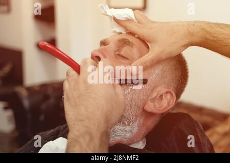 Rasieren mit einem Rasiermesser in einem Friseurladen. Ein bärtiger alter Mann, der in einem Friseurladen rasiert wird. Classic Shave von Stainless Steel Straight Edge Razor. Stockfoto