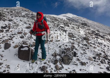 Weibliche Bergsteigertouristin wandert auf gefrorenen Felsen und Steinen über Wolken in großen Bergen vor der Eiskappe auf dem Gipfel des Mount Ararat Stockfoto