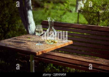 Glas und Dekanter mit Weißwein auf einem Tisch im Garten. Ein Frühlingsabend inmitten der Natur. Vorderansicht. Stockfoto