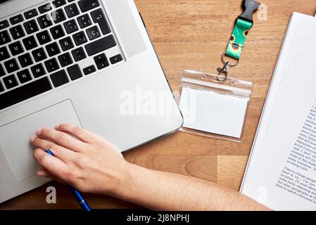 Ansicht von oben auf einen Schreibtisch, eine unbekannte Person, die studiert oder arbeitet, einen Laptop, bedruckte Blätter und einen leeren Personalausweis mit einem Trageband aus Sonnenblumen, Symbol für p Stockfoto