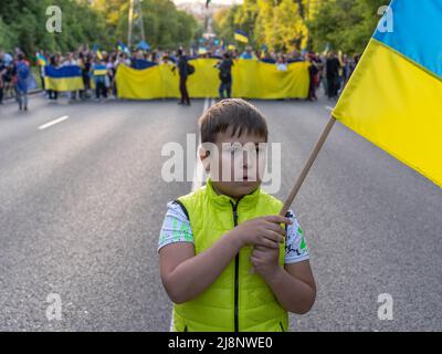 Sofia, Bulgarien - 09. Mai 2022: Ein Kind mit ukrainischer Flagge in den Händen steht vor einer Reihe von Menschen, die mit einer riesigen Flagge der Ukraine in die Stadt gehen Stockfoto