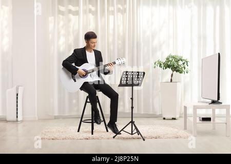 Ein junger Mann in einem schwarzen Anzug, der eine akustische Gitarre spielt und auf einem Stuhl in einem Raum zu Hause sitzt Stockfoto
