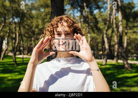 Porträt einer jungen Rotschopf Frau bedeckt ihren Mund mit transparenten Smartphone-Bildschirm über grünen Stadtpark Hintergrund, im Freien. Lächelnd am Telefon, di Stockfoto