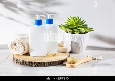 Ein Spender mit Shampoo und Haarspülung, zwei Stücke weißer Seife, eine natürliche Borstenbürste und ein aufgerolltes Gesichtstuch befinden sich auf einem Holzbecher, echiv Stockfoto