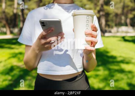 Sportliche Frau mit weißem T-Shirt und schwarzer Yoga-Hose, die Smartphone und eine Tasse Kaffee im Stadtpark im Freien hält. Verwenden des Telefons oder Scrollen in sozialen Netzwerken Stockfoto