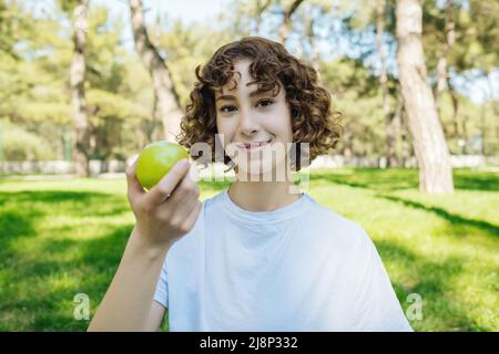 Junge glückliche Rothaarige Frau mit weißem T-Shirt und grünem Apfel und Blick auf die Kamera im Stadtpark, im Freien. Bio und gesunde Ernährung Konzept. Gr Stockfoto